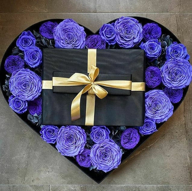 Heart-shaped box filled with purple roses and a black gift box with a gold ribbon on a stone surface.