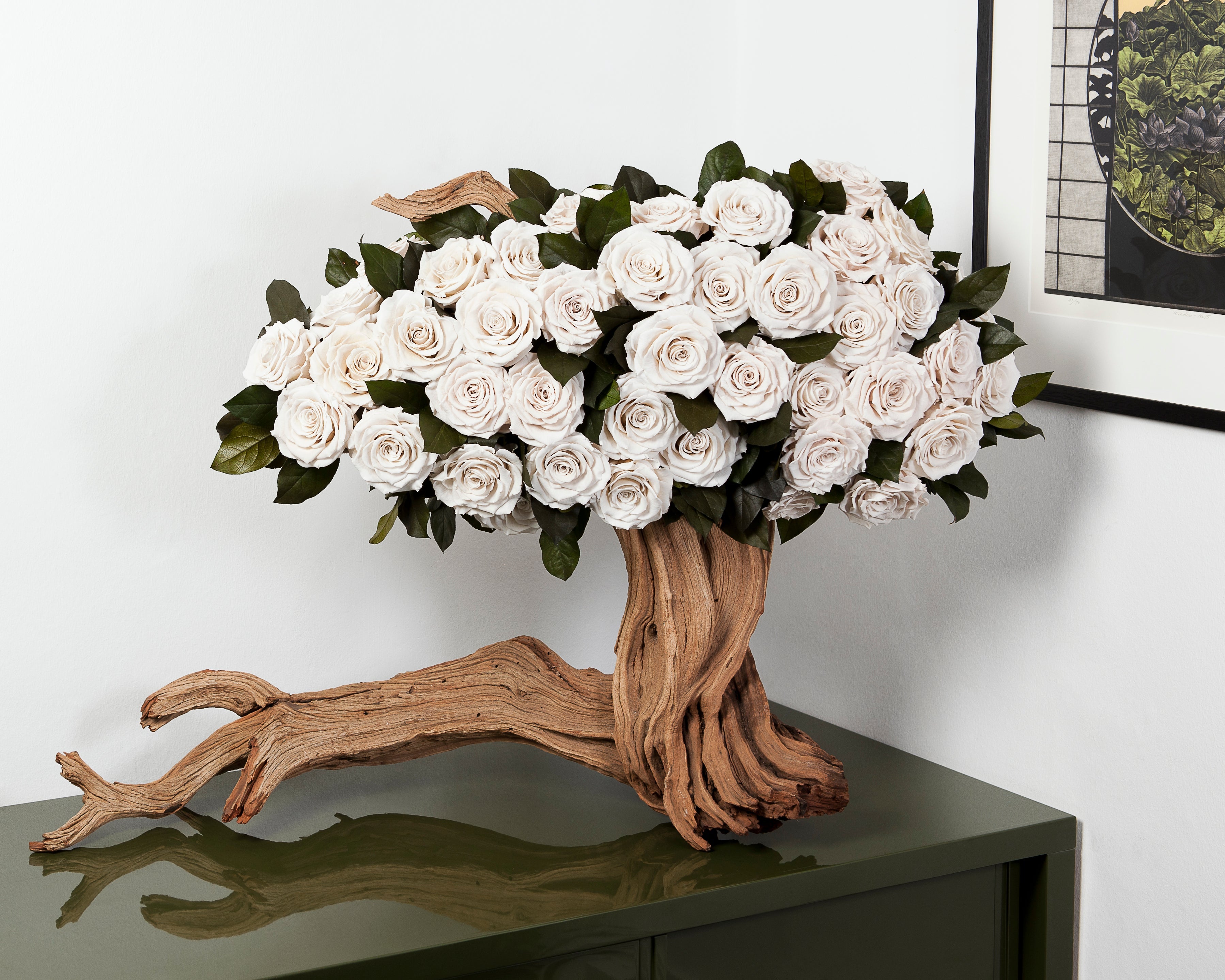 Decorative arrangement of white roses on a wooden base resembling a tree, placed on a reflective surface with a neutral background.